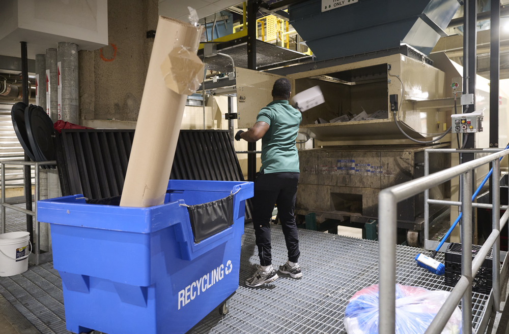 An environmental services worker throws a cardboard item into a recycling compressor at a loading dock.